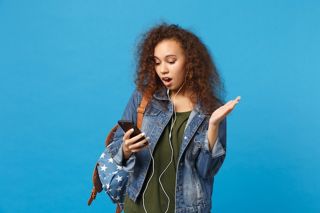 Jonge Afro-Amerikaanse meisje tiener student in denim kleding, rugzak koptelefoon geïsoleerd op blauwe muur
