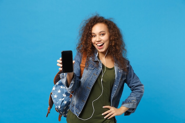 Jonge Afro-Amerikaanse meisje tiener student in denim kleding, rugzak koptelefoon geïsoleerd op blauwe muur