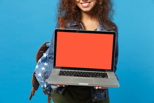 Jonge Afro-Amerikaanse meisje tiener student in denim kleding, rugzak werk op pc geïsoleerd op blauwe muur