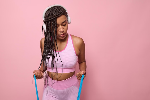 Jonge Afro-Amerikaanse sportieve vrouw in roze trainingspak, concentreer je op het doen van oefeningen op armen met elastische weerstand fitnessband, geïsoleerd op roze achtergrond met kopieerruimte