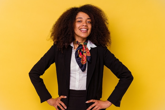 Jonge Afro-Amerikaanse stewardess geïsoleerd op gele achtergrond vertrouwen houden handen op de heupen.