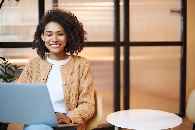 Jonge Afro-Amerikaanse succesvolle mooie vrouw die op laptop werkt en glimlachend naar de camera kijkt