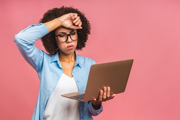 Jonge afro-amerikaanse vermoeide ernstige trieste zakenvrouw met krullend haar die laptop gebruikt die over roze achtergrond wordt geïsoleerd