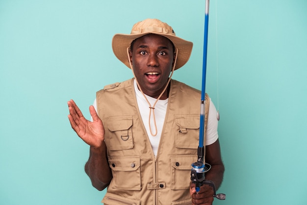 Jonge Afro-Amerikaanse visser met staaf geïsoleerd op blauwe achtergrond verrast en geschokt.