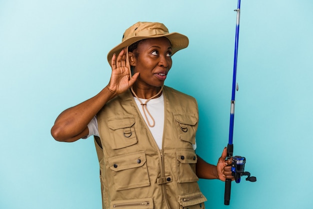 Jonge Afro-Amerikaanse vissersvrouw die een staaf vasthoudt die op een blauwe achtergrond wordt geïsoleerd en die een roddel probeert te luisteren.