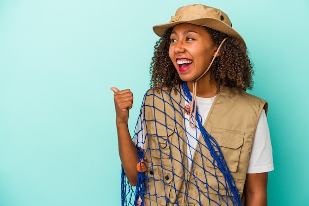 Jonge afro-amerikaanse vissersvrouw met net geïsoleerd op blauwe achtergrond wijst met duimvinger weg, lachend en zorgeloos.