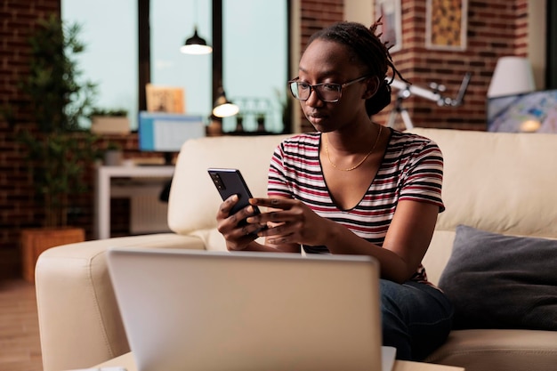 Jonge afro-amerikaanse vrouw bericht aan het typen op smartphone, browsen op sociale media, chatten. Freelancer met pauze, ontspannen student in glazen surfen op internet op mobiele telefoon