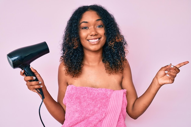 Jonge afro-amerikaanse vrouw die een douchehanddoek draagt en een droger vasthoudt glimlachend blij wijzend met hand en vinger naar de zijkant