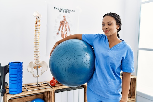 Jonge afro-amerikaanse vrouw die een uniforme fysiotherapeut draagt die een fitte bal vasthoudt in de kliniek