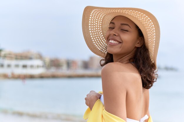 Jonge afro-amerikaanse vrouw die lacht zelfverzekerd met zomerhoed en bikini op het strand