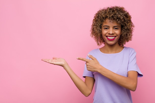 Jonge Afro-Amerikaanse vrouw die lacht, zich gelukkig, zorgeloos en tevreden voelt, wijzend op concept of idee op kopie ruimte aan de zijkant tegen roze muur
