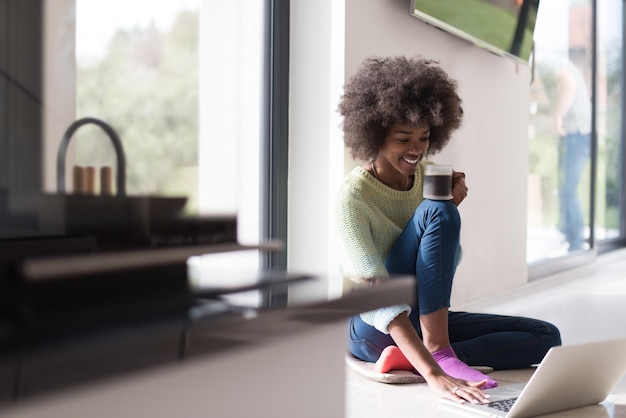 Jonge afro-amerikaanse vrouw die lacht zittend op de vloer bij een helder raam terwijl ze naar een opengeklapte laptop kijkt en thuis een mok vasthoudt