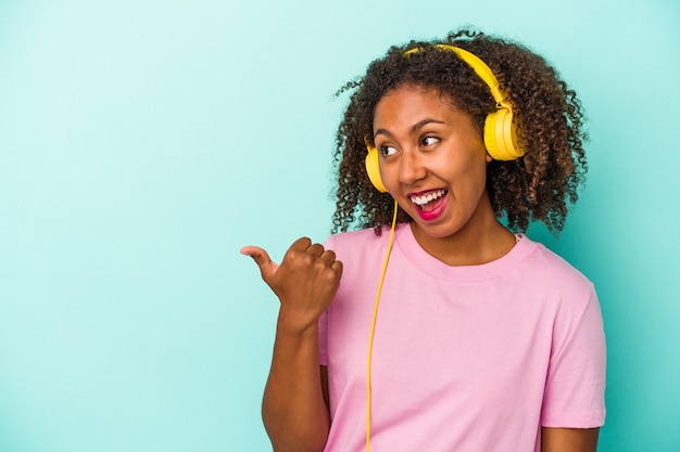 Jonge afro-amerikaanse vrouw die naar muziek luistert geïsoleerd op blauwe achtergrond wijst met duimvinger weg, lachend en zorgeloos.