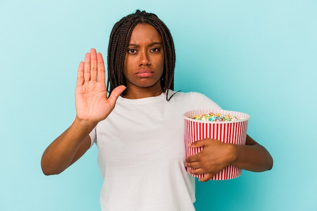 Jonge afro-amerikaanse vrouw die pop likdoorns eet geïsoleerd op een blauwe achtergrond die staat met uitgestrekte hand die een stopbord toont, waardoor je wordt verhinderd.