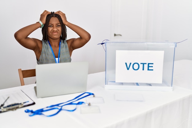 Jonge afro-amerikaanse vrouw die werkt bij politieke verkiezingen die bij de stemming zitten en lijden aan hoofdpijn, wanhopig en gestrest vanwege pijn en migraine. handen op het hoofd.
