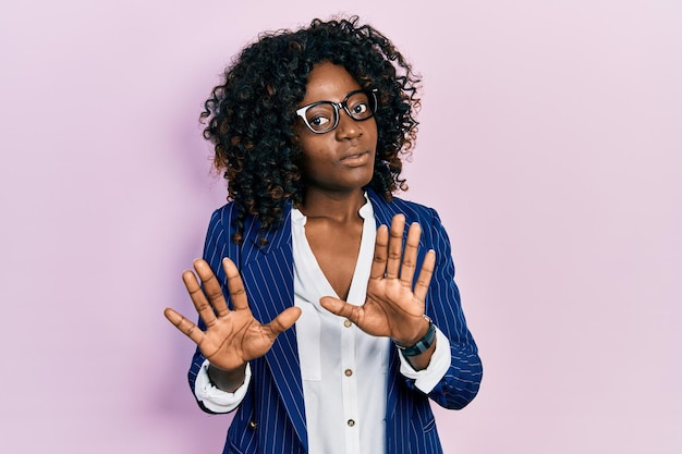 Jonge afro-amerikaanse vrouw die zakelijke kleding en een bril draagt die de handpalmen wegtrekt en weigering en ontkenning toont met een bange en walgelijke uitdrukking, stop en verboden