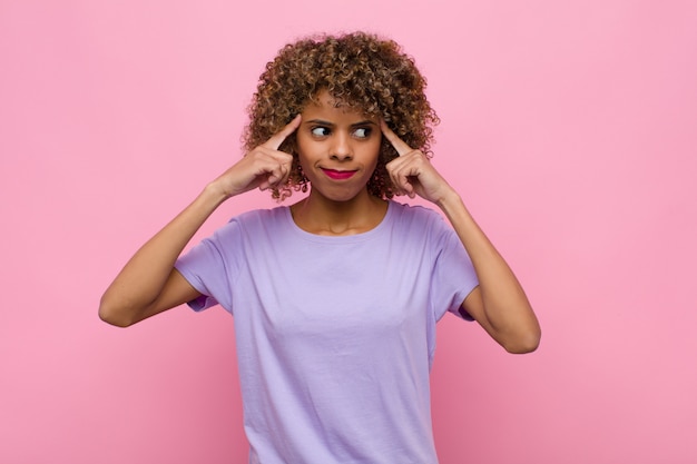 Jonge afro-amerikaanse vrouw die zich verward of twijfelt, zich concentreert op een idee, hard nadenkt, ruimte aan de zijkant op roze muur wil kopiëren