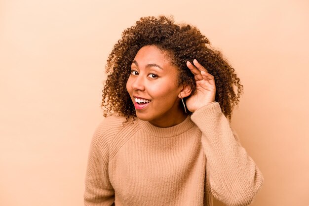 Jonge Afro-Amerikaanse vrouw geïsoleerd op beige achtergrond die een roddel probeert te luisteren