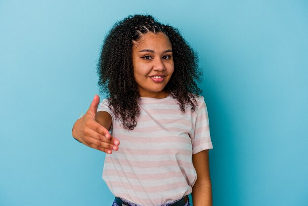 Jonge afro-amerikaanse vrouw geïsoleerd op blauwe achtergrond hand uitrekken op camera in begroeting gebaar.