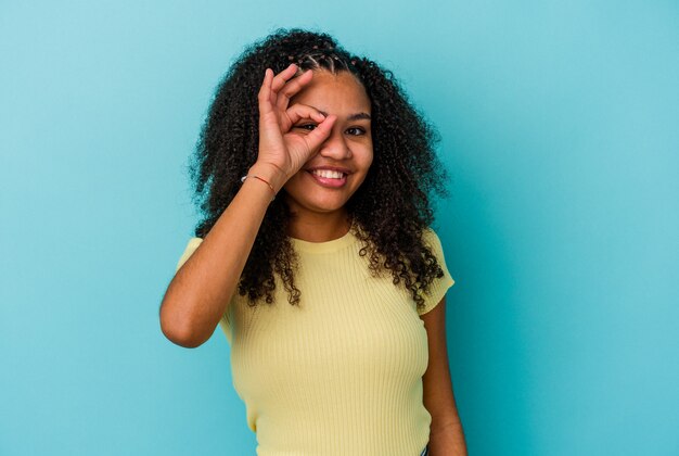 Jonge Afro-Amerikaanse vrouw geïsoleerd op blauwe achtergrond opgewonden houden ok gebaar op oog.