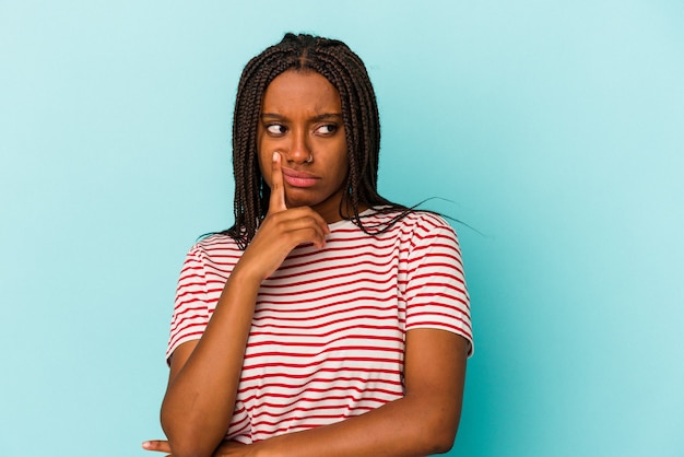 Jonge afro-amerikaanse vrouw geïsoleerd op blauwe achtergrond zijwaarts kijkend met twijfelachtige en sceptische uitdrukking.