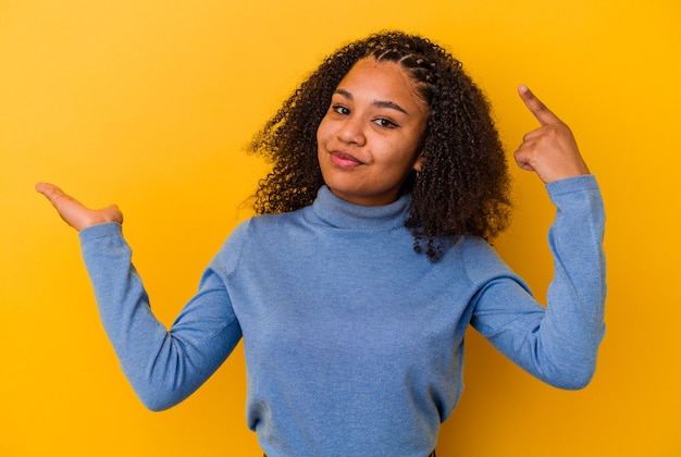 Jonge afro-amerikaanse vrouw geïsoleerd op een gele achtergrond die een product bij de hand houdt en toont.