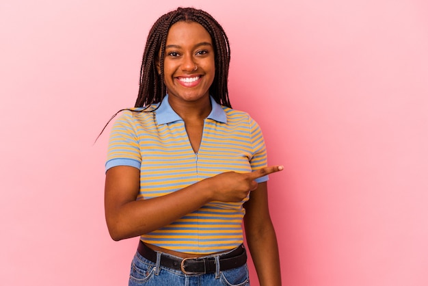 Jonge afro-amerikaanse vrouw geïsoleerd op roze achtergrond glimlachend en opzij wijzend, iets tonend op lege ruimte.
