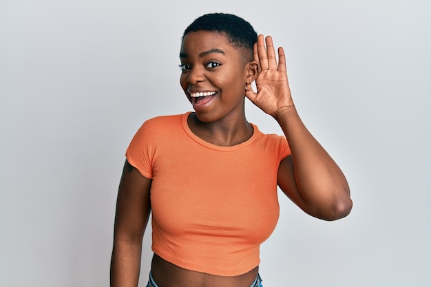 Jonge afro-amerikaanse vrouw, gekleed in casual oranje t-shirt glimlachend met hand over oor luisterend naar een hoorzitting naar geruchten of roddels doofheid concept