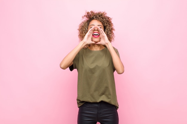 Jonge Afro-Amerikaanse vrouw gelukkig, opgewonden en positief gevoel, het geven van een grote schreeuw met handen naast de mond, uitroepen tegen roze muur