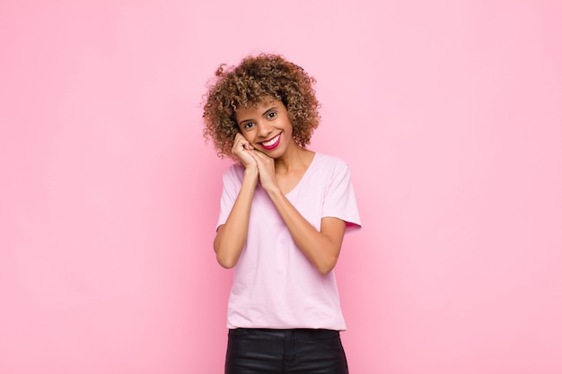 Jonge Afro-Amerikaanse vrouw gevoel in liefde en op zoek schattig, schattig en gelukkig, romantisch lachend met handen naast gezicht tegen roze muur