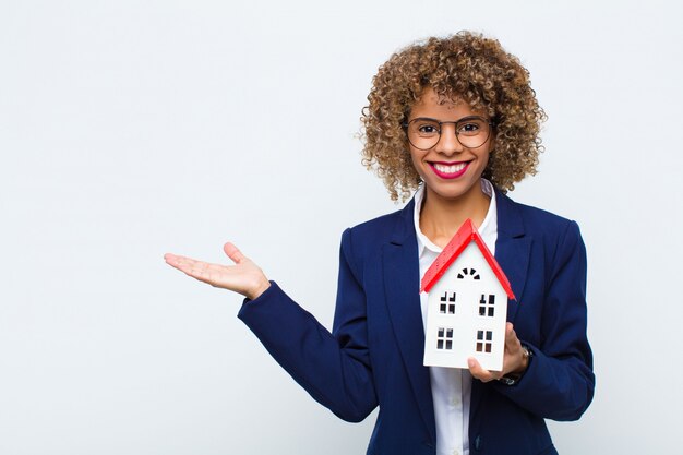 Jonge Afro-Amerikaanse vrouw lacht vrolijk, voelt zich gelukkig en toont een concept in kopie ruimte met palm van de hand met huis model