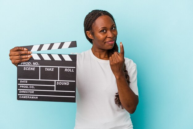 Jonge afro-amerikaanse vrouw met een filmklapper geïsoleerd op een blauwe achtergrond, wijzend met de vinger naar je alsof uitnodigend dichterbij komt.