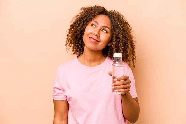 Jonge Afro-Amerikaanse vrouw met een fles water geïsoleerd op een beige achtergrond, dromend van het bereiken van doelen en doeleinden