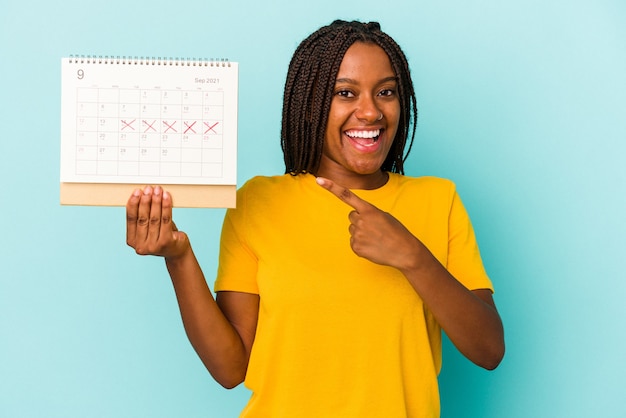 Jonge afro-amerikaanse vrouw met een kalender geïsoleerd op een blauwe achtergrond glimlachend en opzij wijzend, iets tonend op de lege ruimte.