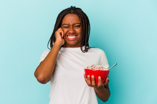 Jonge afro-amerikaanse vrouw met een kom cornflakes geïsoleerd op een blauwe achtergrond die oren bedekt met handen.