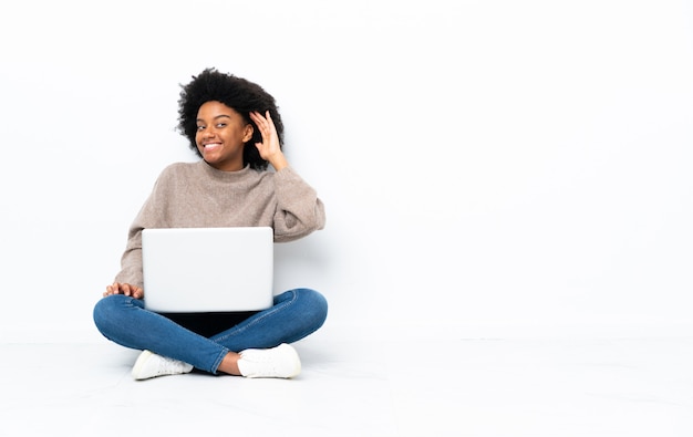 Jonge afro-amerikaanse vrouw met een laptop zittend op de vloer luisteren naar iets door hand op het oor te zetten