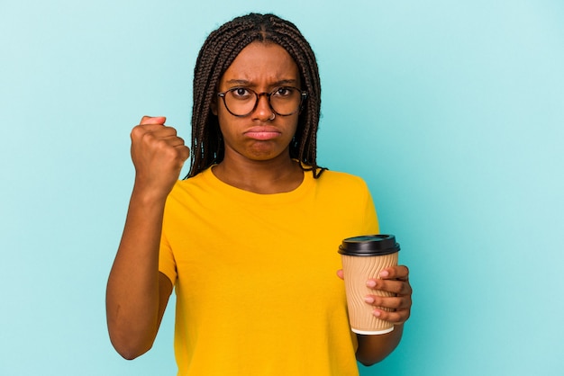 Jonge Afro-Amerikaanse vrouw met een take-away koffie geïsoleerd op een blauwe achtergrond met vuist naar camera, agressieve gezichtsuitdrukking.