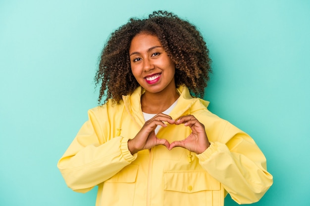 Jonge afro-amerikaanse vrouw met krullend haar geïsoleerd op blauwe achtergrond glimlachend en met een hartvorm met handen.