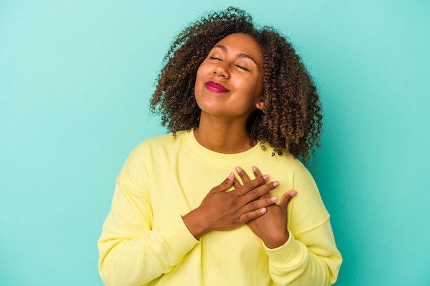 Jonge afro-amerikaanse vrouw met krullend haar geïsoleerd op blauwe achtergrond heeft vriendelijke uitdrukking, palm op borst drukken. Liefdesconcept.