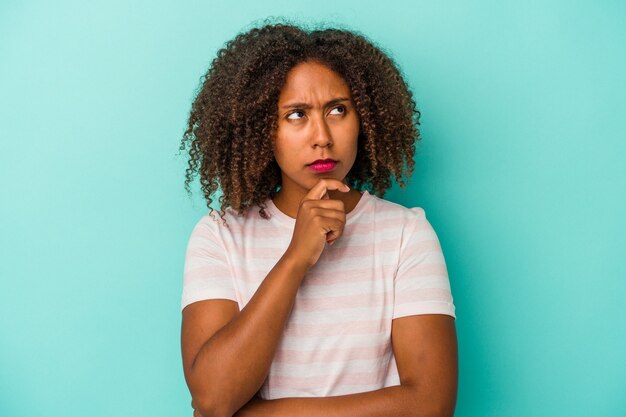 Jonge Afro-Amerikaanse vrouw met krullend haar geïsoleerd op blauwe achtergrond zijwaarts kijkend met twijfelachtige en sceptische uitdrukking.