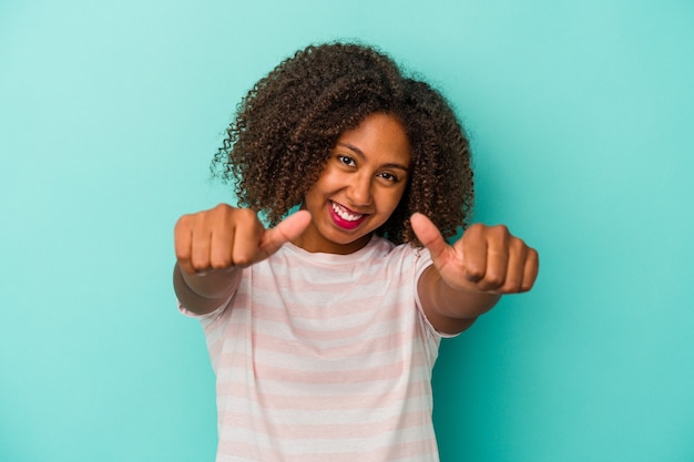 Jonge afro-amerikaanse vrouw met krullend haar geïsoleerd op een blauwe achtergrond die beide duimen opheft, glimlachend en zelfverzekerd.