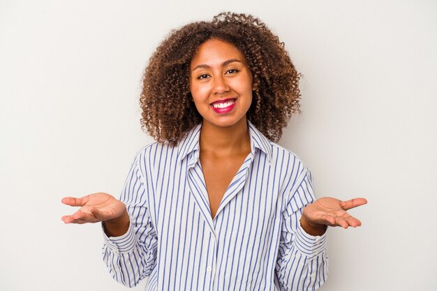 Jonge afro-amerikaanse vrouw met krullend haar geïsoleerd op een witte achtergrond maakt schaal met armen, voelt zich gelukkig en zelfverzekerd.