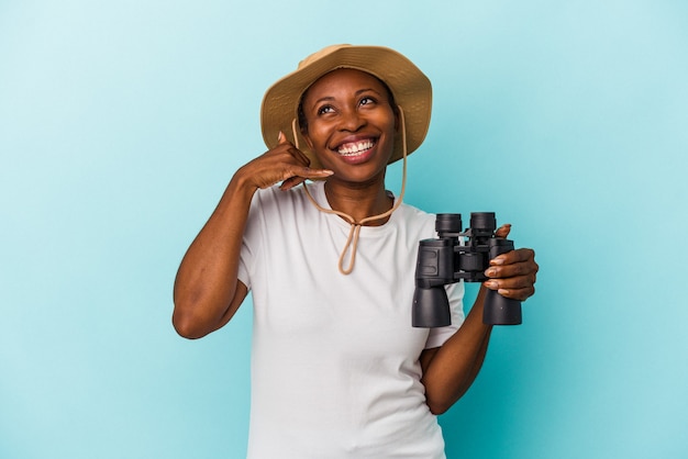 Jonge Afro-Amerikaanse vrouw met verrekijker geïsoleerd op blauwe achtergrond met een mobiel telefoongesprek gebaar met vingers.