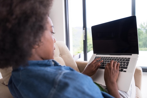 Foto jonge afro-amerikaanse vrouw thuis in de stoel ontspannen in haar luxe woonkamer lezen laptop pc surfen op internet en werken