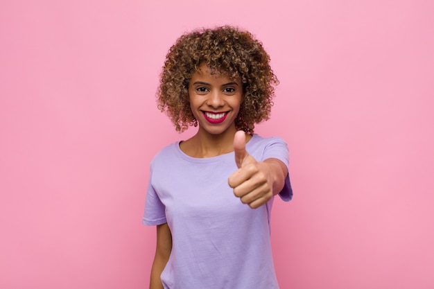 Jonge Afro-Amerikaanse vrouw trots, zorgeloos, zelfverzekerd en gelukkig gevoel, positief glimlachend met duimen tegen roze muur