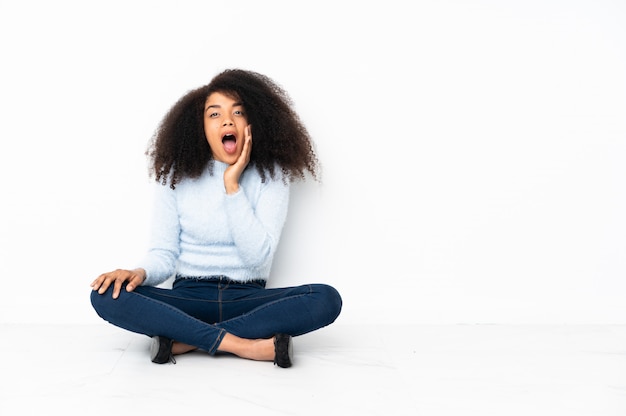 Jonge afro-amerikaanse vrouw zittend op de vloer met verrassing en geschokte gelaatsuitdrukking