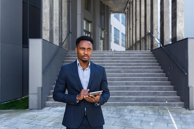 Jonge Afro-Amerikaanse zakenman in formeel pak staande werken met tablet in handen op achtergrond modern kantoorgebouw buiten. Man met smartphone of mobiele telefoon buiten stadsstraat