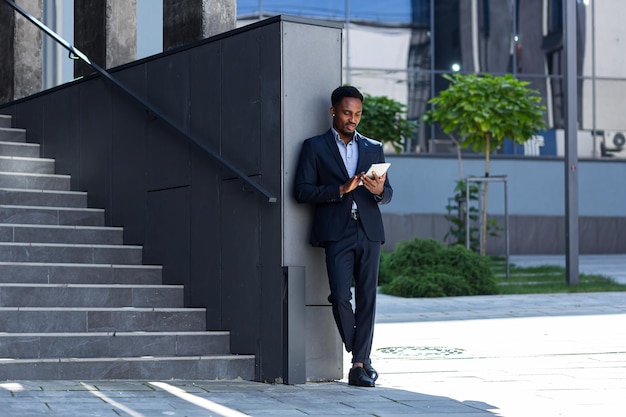 Jonge Afro-Amerikaanse zakenman in formeel pak staande werken met tablet in handen op achtergrond modern kantoorgebouw buiten. Man met smartphone of mobiele telefoon buiten stadsstraat