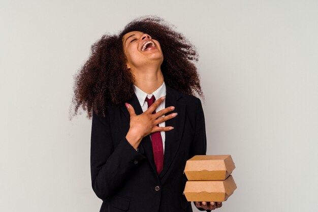 Jonge afro-amerikaanse zakenvrouw met een hamburger geïsoleerd op wit lacht hardop terwijl ze de hand op de borst houdt.