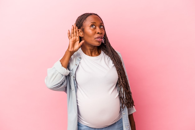 Jonge afro-amerikaanse zwangere vrouw geïsoleerd op roze achtergrond probeert te luisteren naar een roddel.